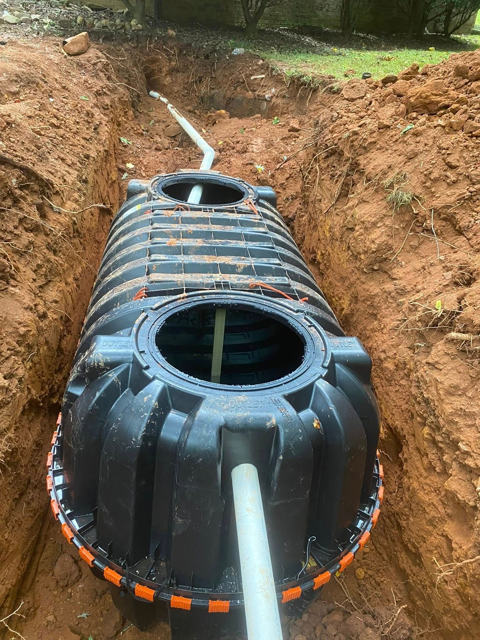 Black septic tank installed in a dirt trench, white pipes connected.