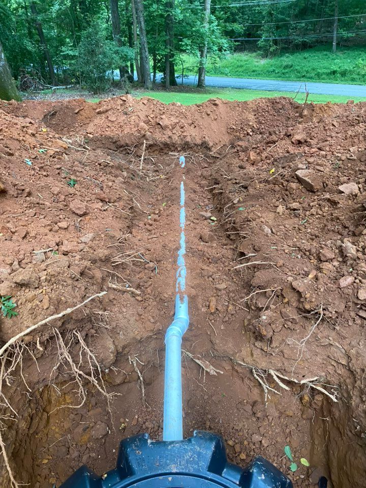 Trench with installed PVC pipe in dirt, leading away from a dark object. Trees and a road are visible in the background.