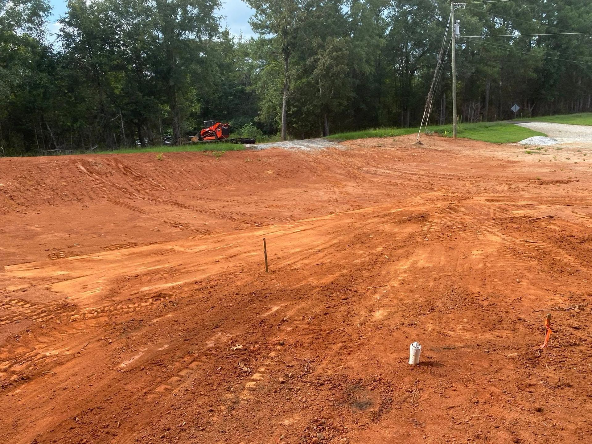 Red dirt construction site with a small tractor and trees in the background.