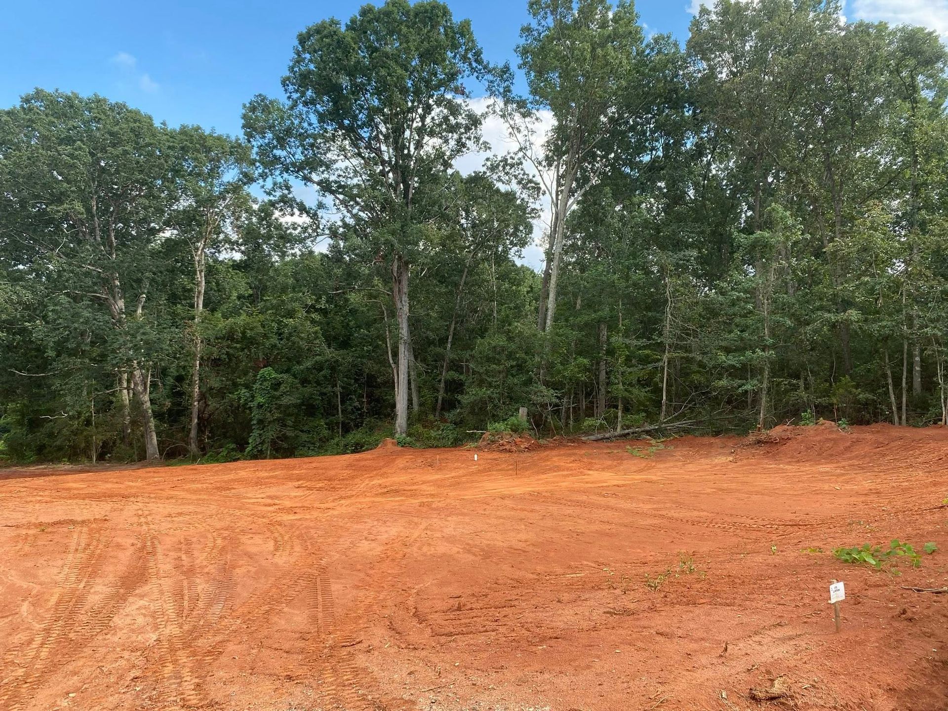 Cleared red dirt lot in front of a line of tall green trees under a blue sky.