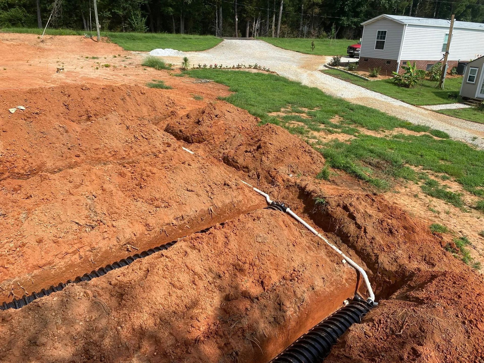Dug trenches with white and black pipes on a dirt lot. A small building is visible in the background.