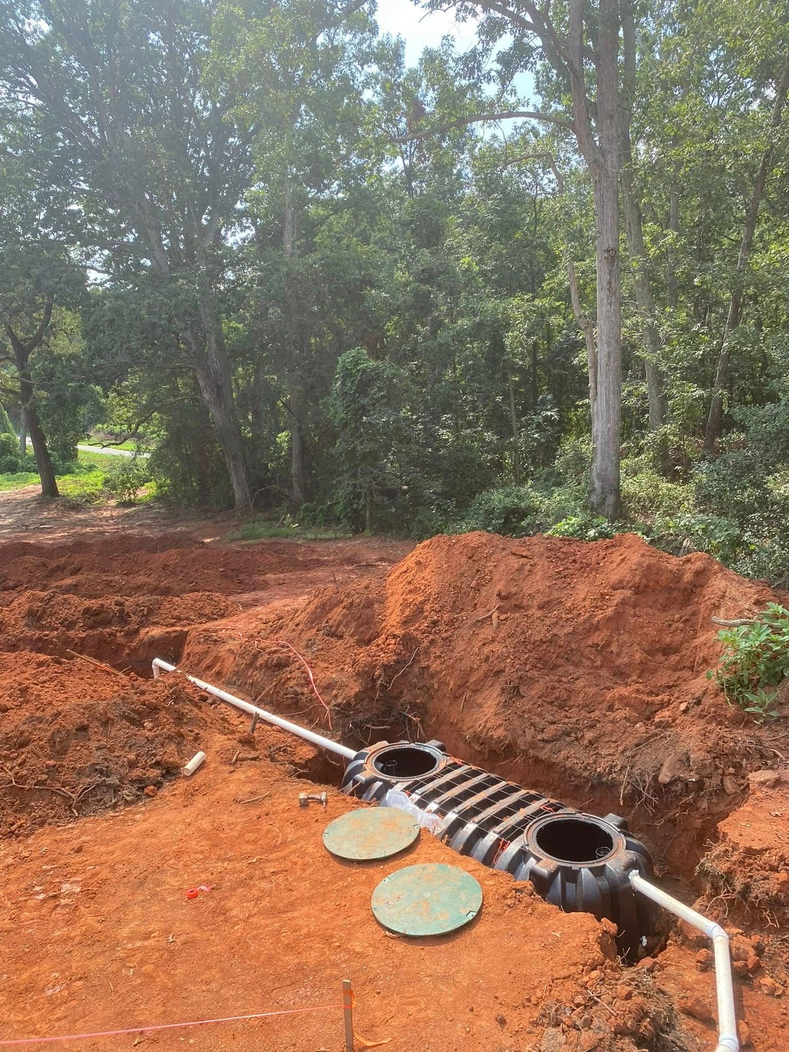 Septic tank installation in progress. Exposed tank with lids, pipes, and red soil. Forest in the background.