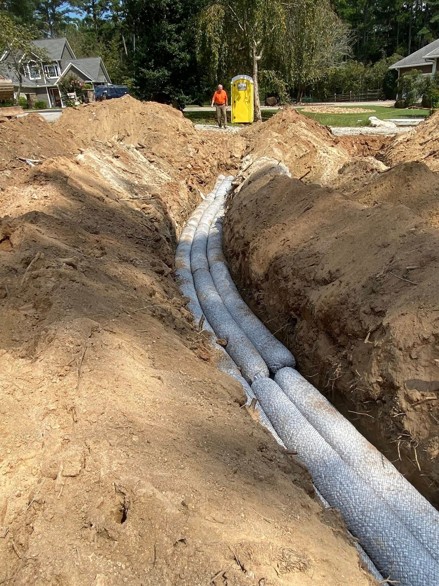 Trench with gray, cylindrical drain pipes. Dirt mounds on either side. A worker stands nearby.
