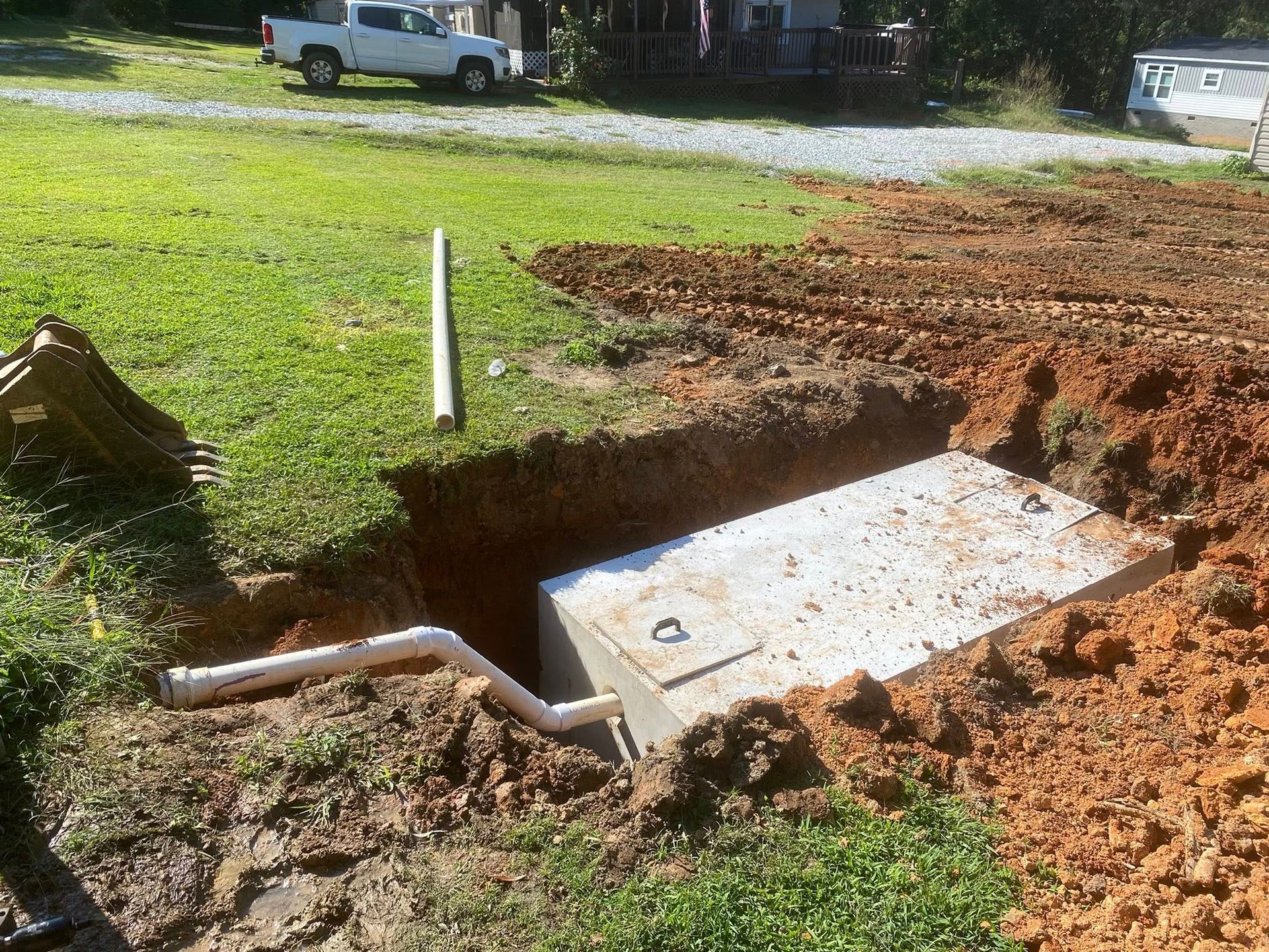 A septic tank installation. A concrete tank is in a dirt excavation with white pipes and a pickup truck in the background.