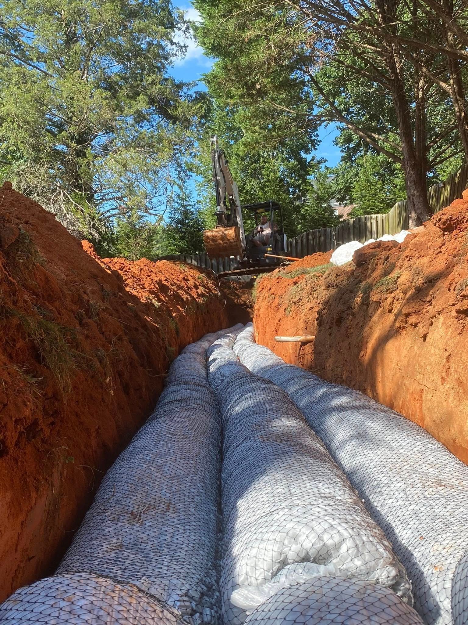 Trench with fabric-wrapped drainage pipes in red clay soil. Trees and blue sky visible in the background.