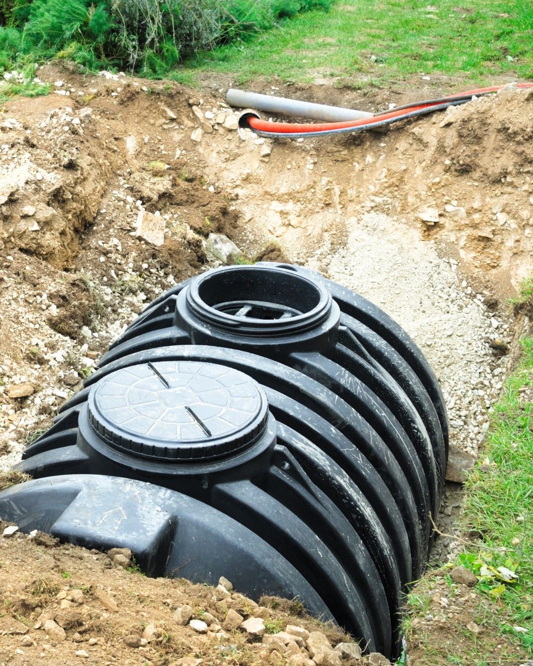 Black ribbed septic tank partially buried in a dirt pit, with piping visible.