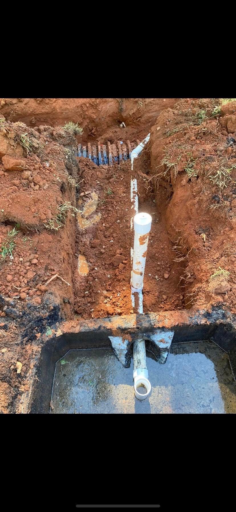A trench in the ground with plumbing pipes and connections exposed, surrounded by dirt and debris.