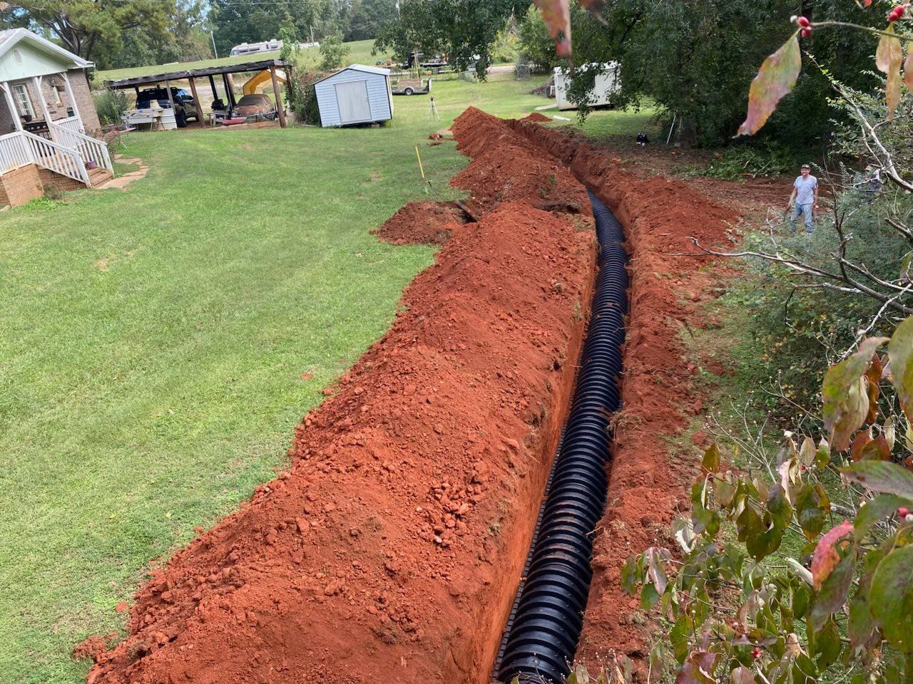 A black corrugated drain pipe in a trench of red soil in a grassy yard.