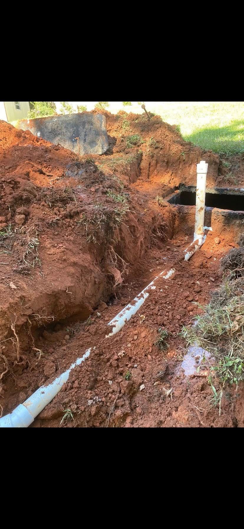 Trench in red soil with white pipes, possibly for plumbing or irrigation in a yard setting.