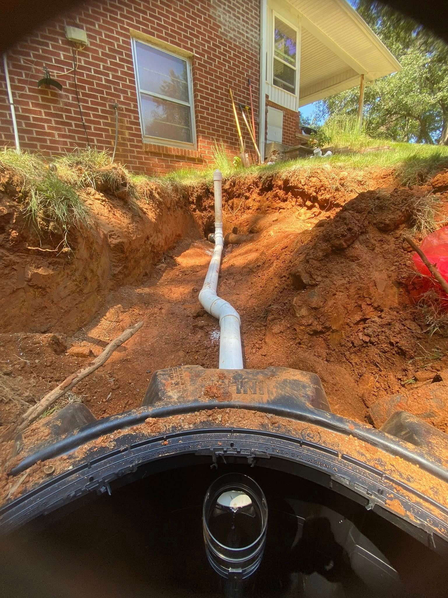 Sump pump installation next to a brick building. White pipe extends from the ground into the excavated hole.