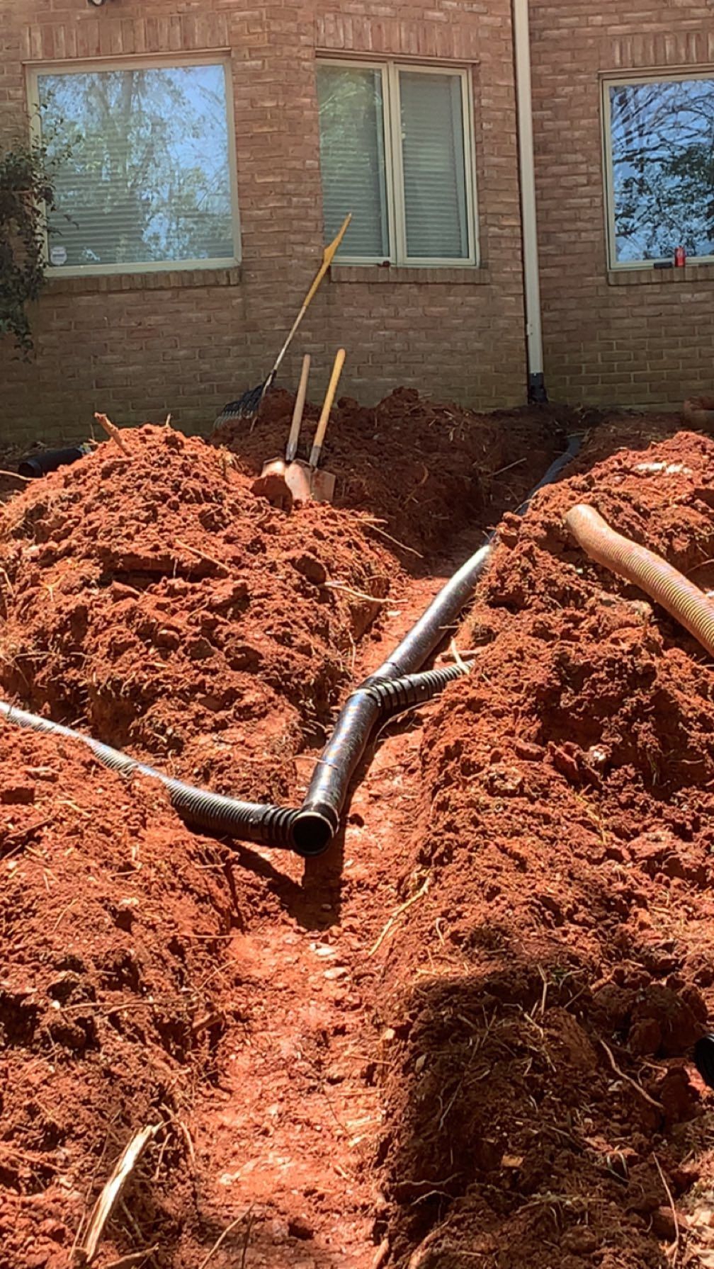 Trench in red dirt with black PVC pipes and yellow stakes, beside a brick building.