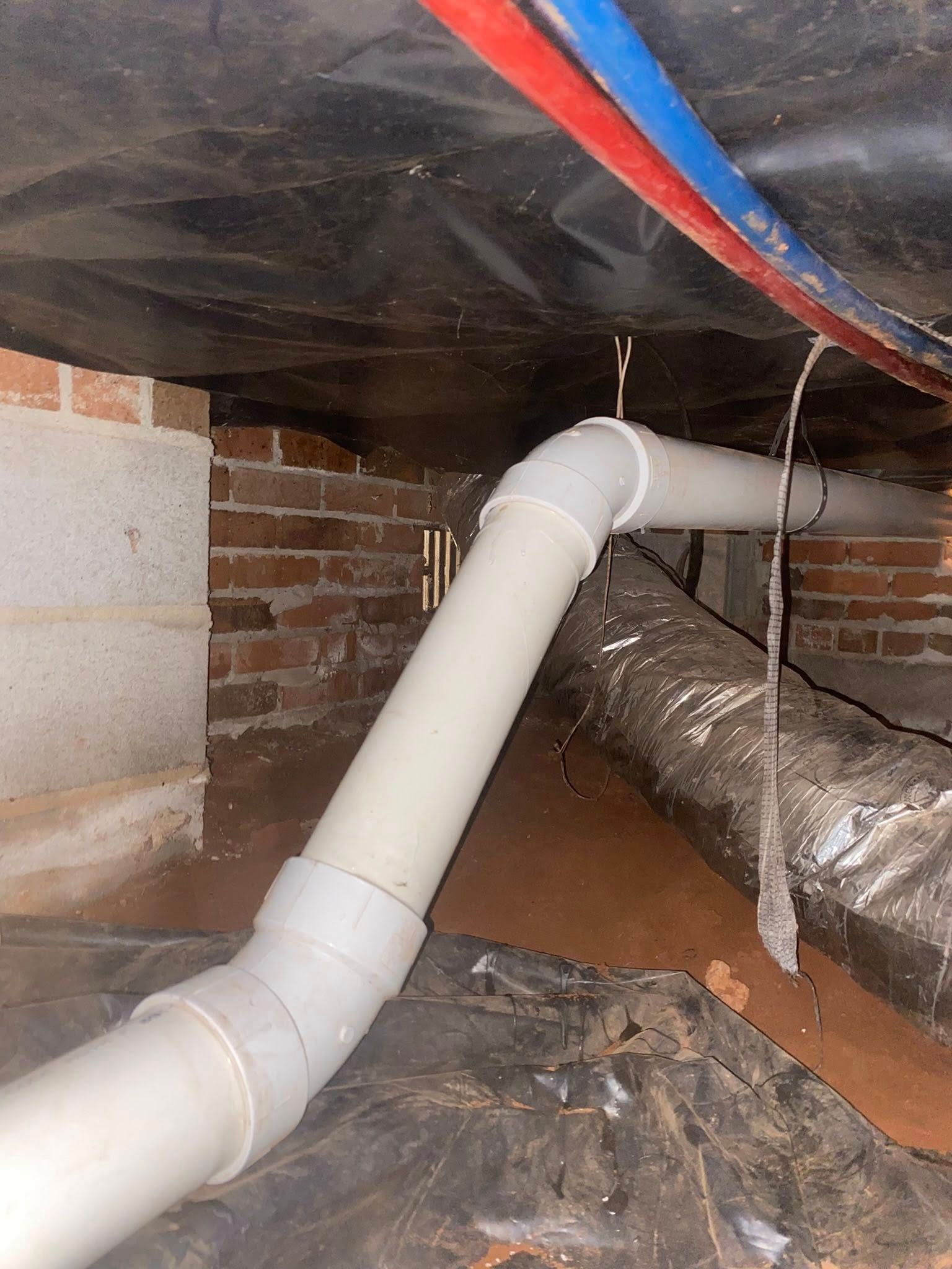 White PVC drain pipe in a crawl space, against brick and dark plastic.