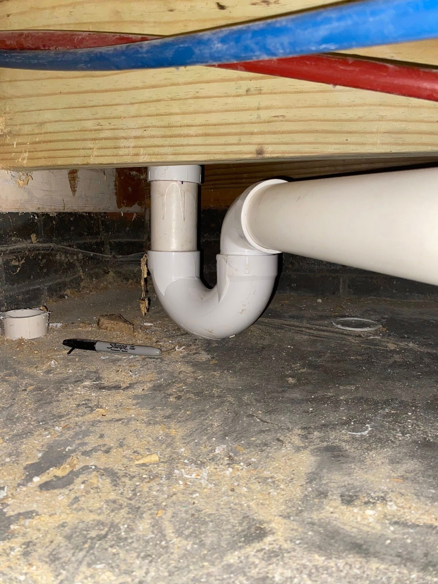 White PVC drain pipe with a P-trap beneath wooden beams, blue and red water pipes above.
