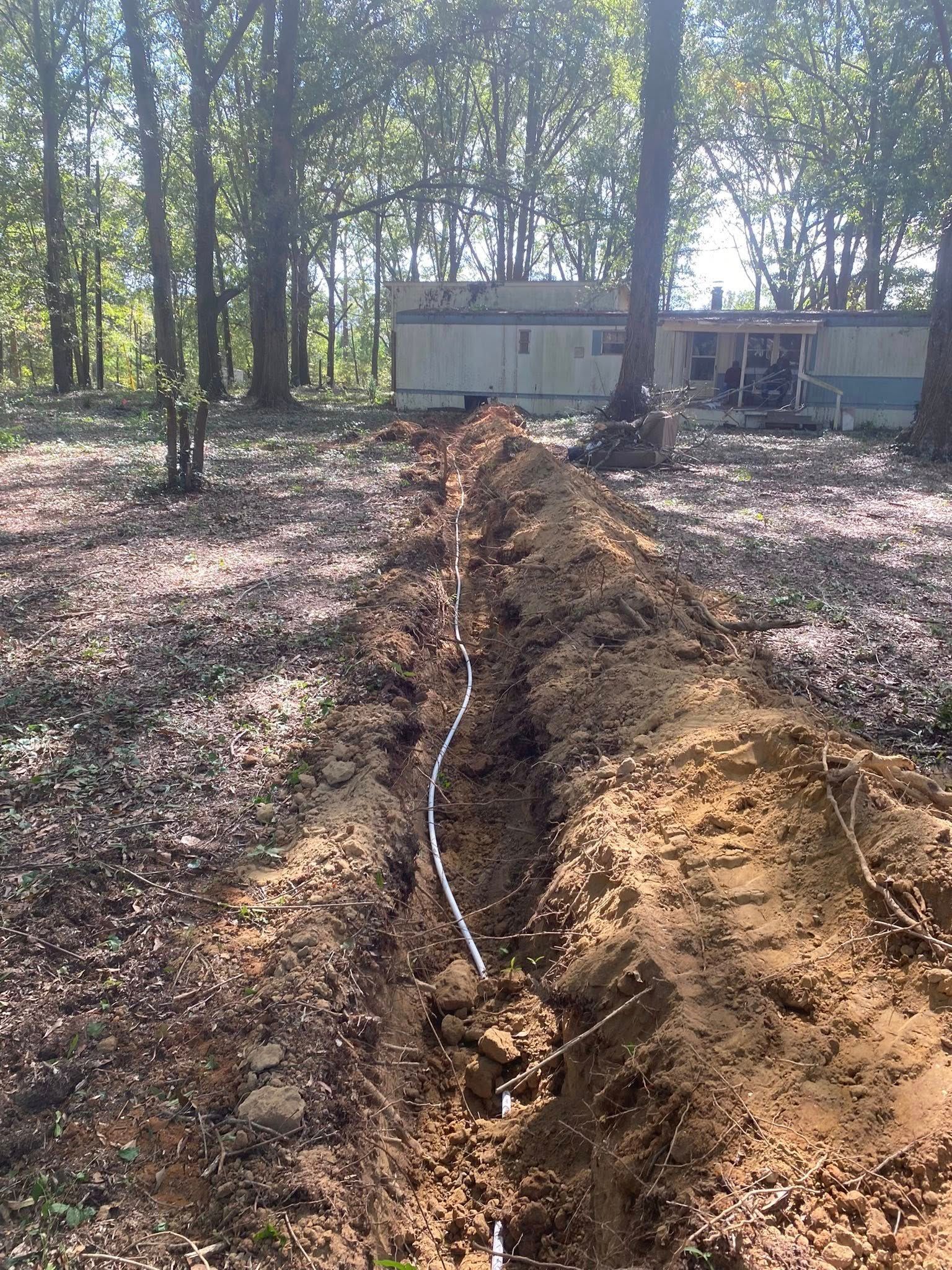 A trench dug in dirt, with a white pipe running through it, leading to a mobile home in a wooded area.