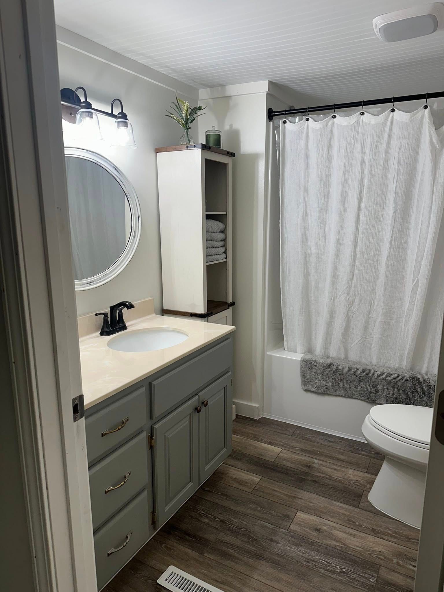 Bathroom with gray vanity, round mirror, white shower curtain, and dark wood-look flooring.