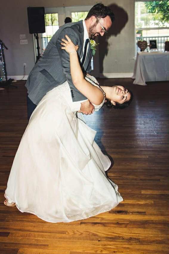 Bride and groom dance, dipping low on a wood floor. The bride wears white, groom in a gray suit.