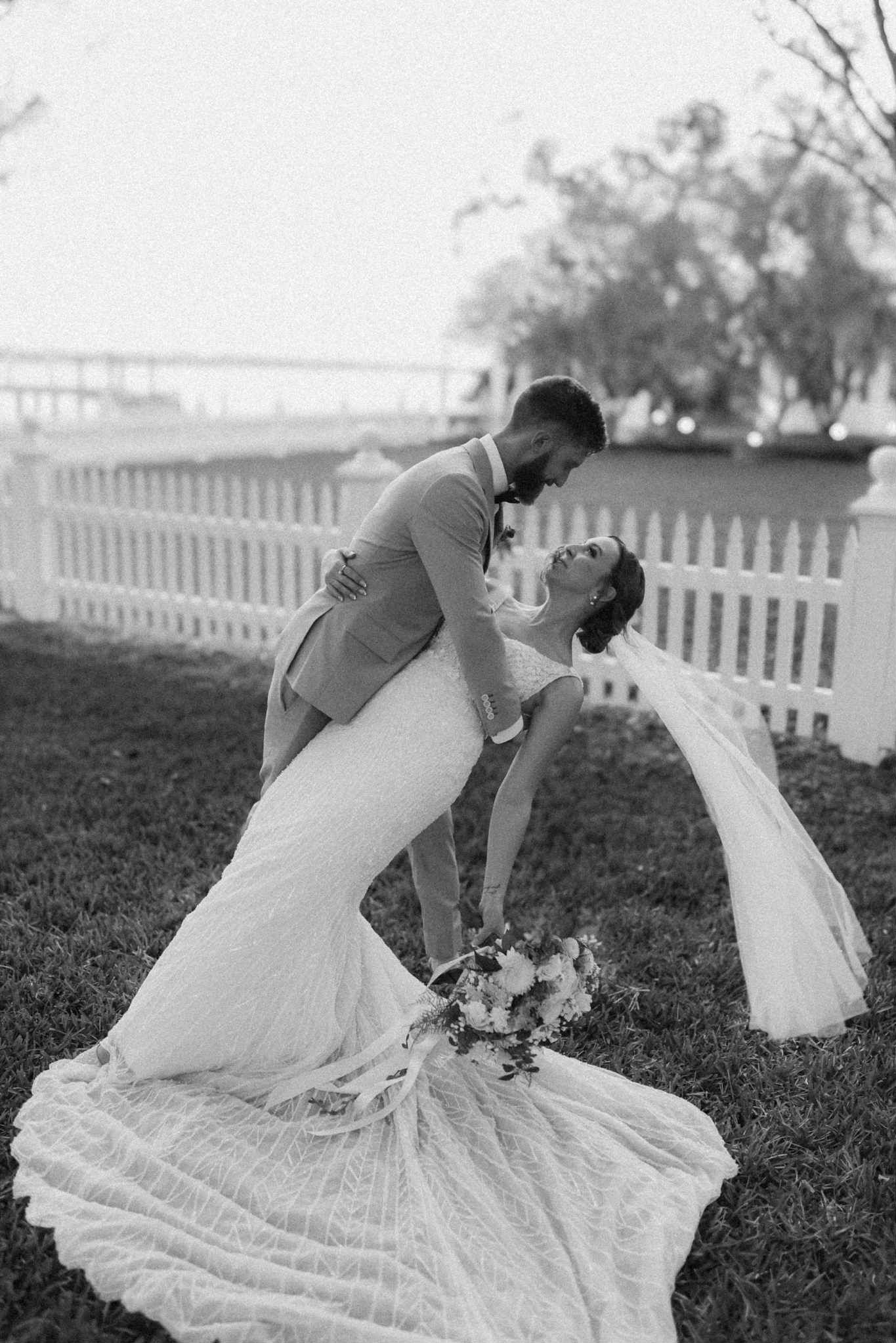 Newlyweds embrace in front of a white picket fence, woman arches backward, veil trailing, man holds her.