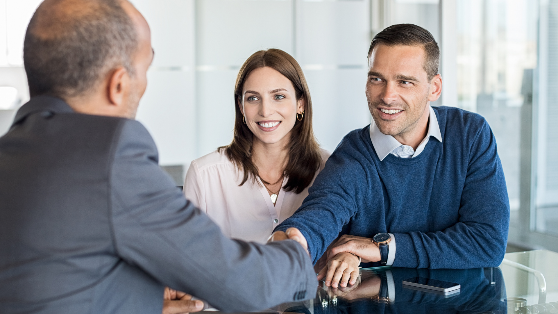 Man in suit shaking hands with a smiling man, with a smiling woman next to him at a table.