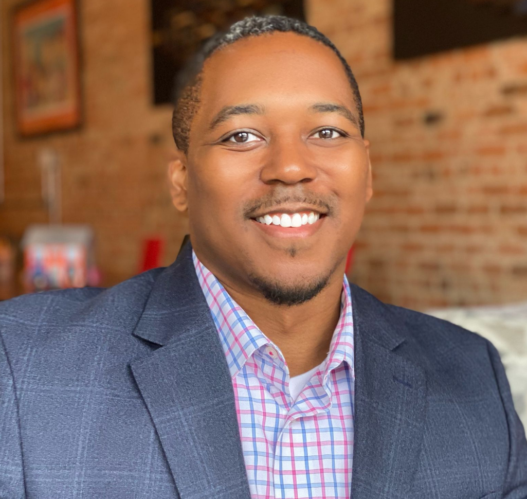 A man wearing a plaid shirt and a vest is standing in front of a tree.