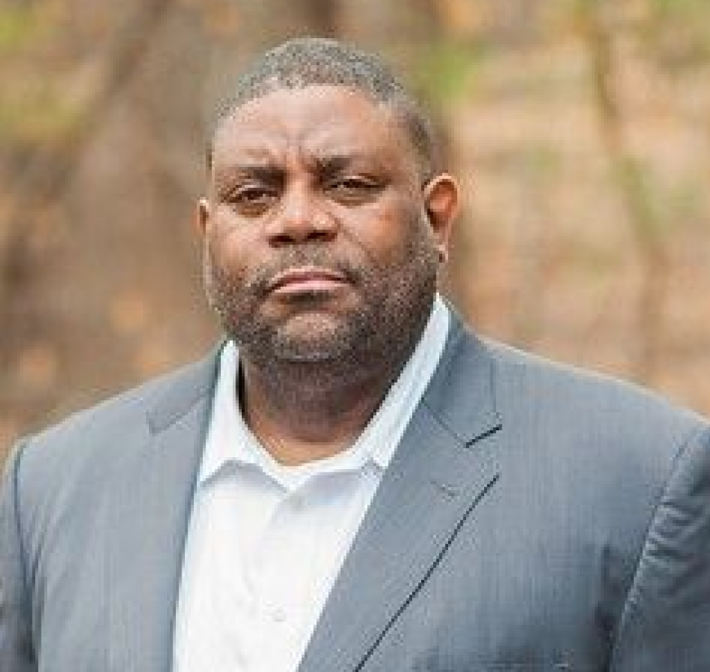 A man wearing a plaid shirt and a vest is standing in front of a tree.