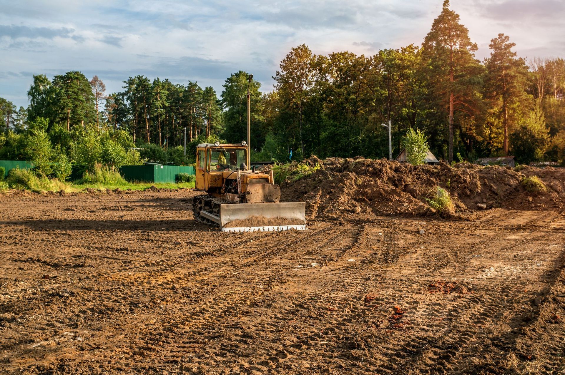 Bulldozer leveling a dirt field beside a tree line under a partly cloudy sky