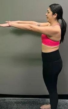 Woman in pink top and black leggings, arms outstretched, standing near a wall.