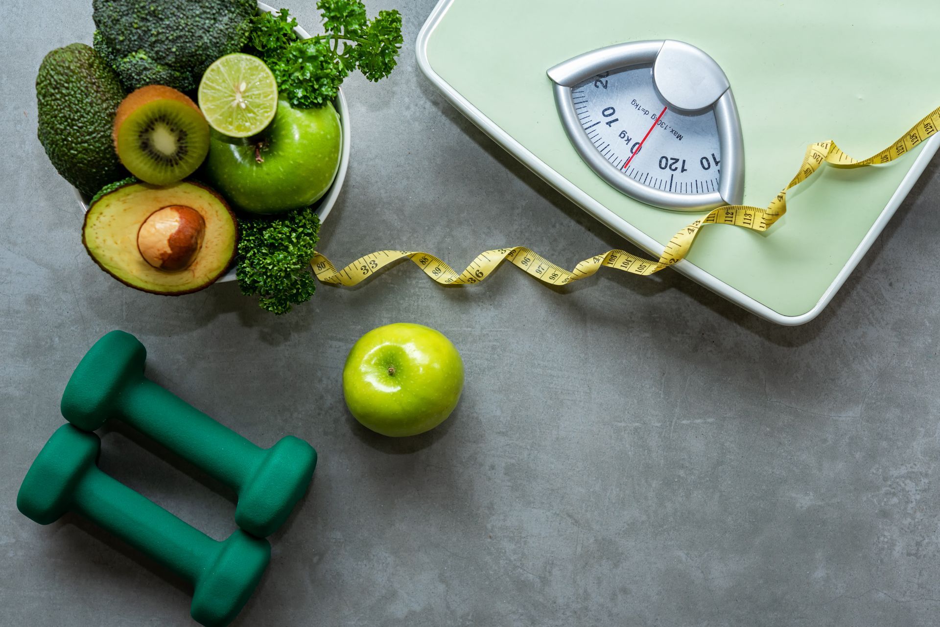 Healthy eating and fitness tools: Bowl of fruit, dumbbells, scale, and measuring tape on a gray surface.