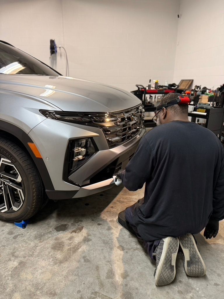 Man in black work clothes kneels, examining the front bumper of a silver SUV in a garage.