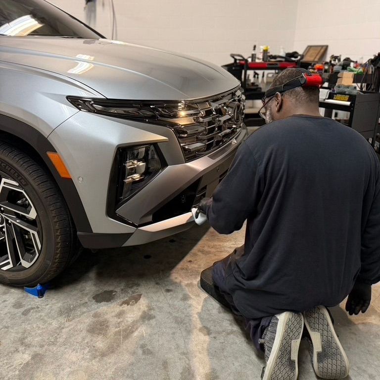A man is kneeling down in front of a silver car in a garage.