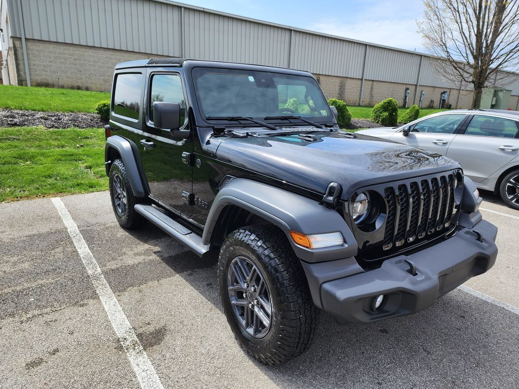 Black Jeep Wrangler parked in a parking lot. Gray trim and wheels, green grass, and a building are visible.