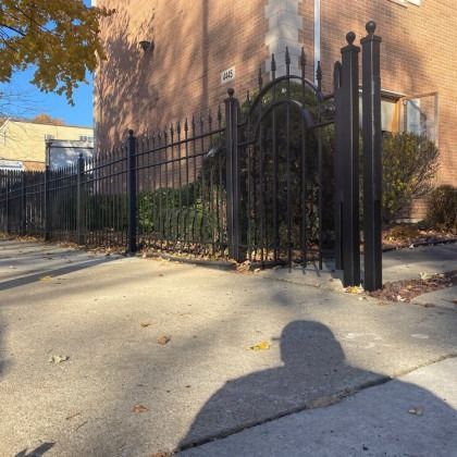 Black metal fence and gate bordering a sidewalk, in front of a brick building.