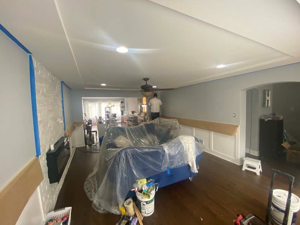 Room being painted: blue tape on walls, plastic-covered furniture, painter working near ceiling fan, hardwood floors.
