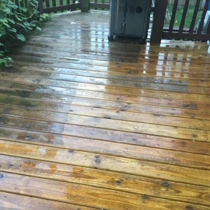 Wet wooden deck, part of which has been cleaned. Some greenery and a grill are visible.