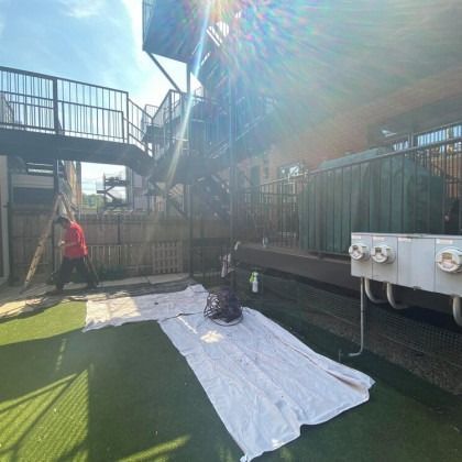 Person sweeping on a green surface with a white sheet. Metal stairs and green building in the background, bright sunlight.
