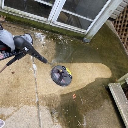 A person using a pressure washer with a circular cleaning head on a concrete patio next to a building.