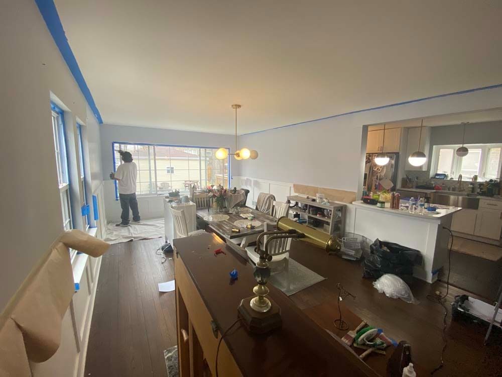 Person paints a light-colored room, windows taped. Table, kitchen, and debris present.