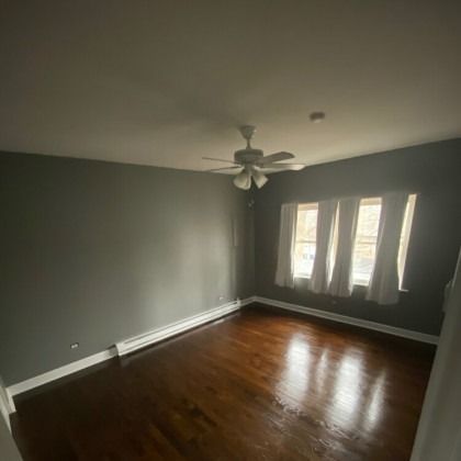 Empty room with dark hardwood floors, gray walls, and a ceiling fan. Three windows with curtains.