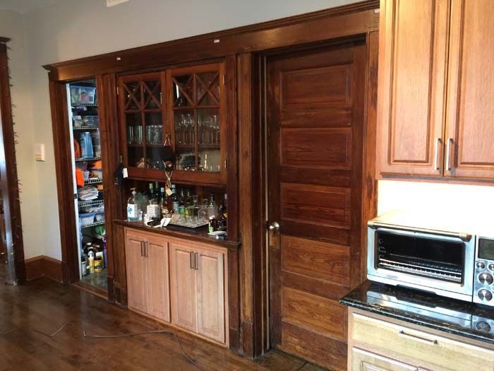 Wooden built-in bar with glass-fronted cabinets, bottles, and a door, next to a pantry and kitchen cabinets.