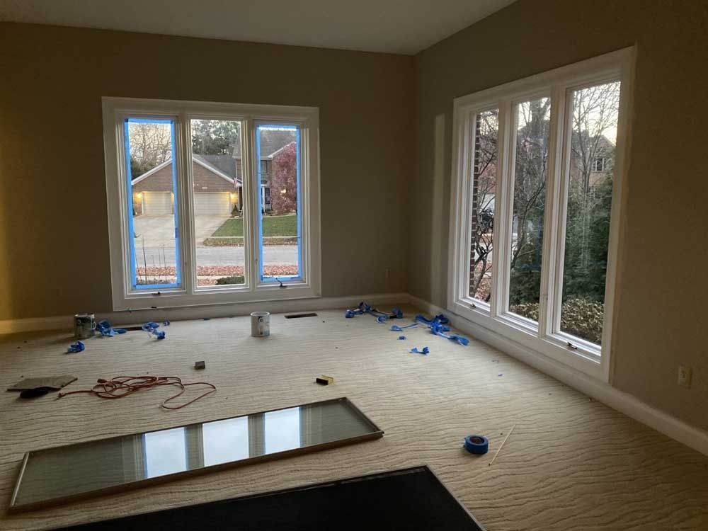 Room with large windows, tan walls, and a light-colored carpet. Debris and a pane of glass are on the floor.