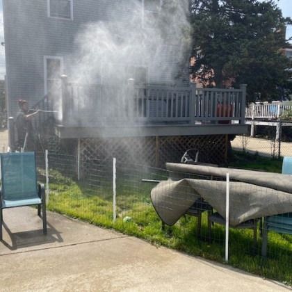 Person spraying mist on a deck with chairs and a small fence in a backyard setting.