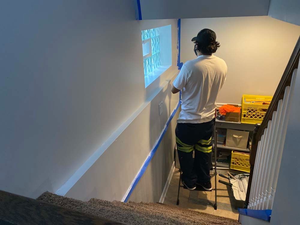 Person taping a wall near a window and stairs; painter wearing a mask, standing on a step ladder.