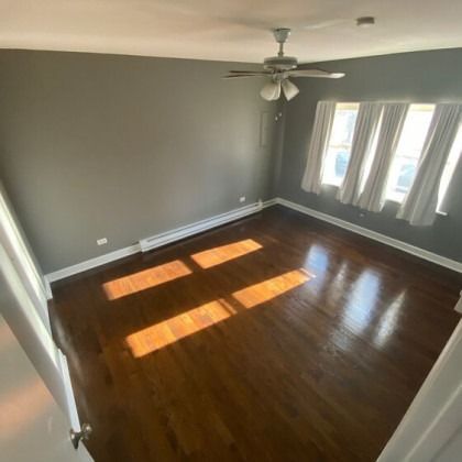 Empty room with dark wood floors, gray walls, three windows with white curtains, and a ceiling fan.