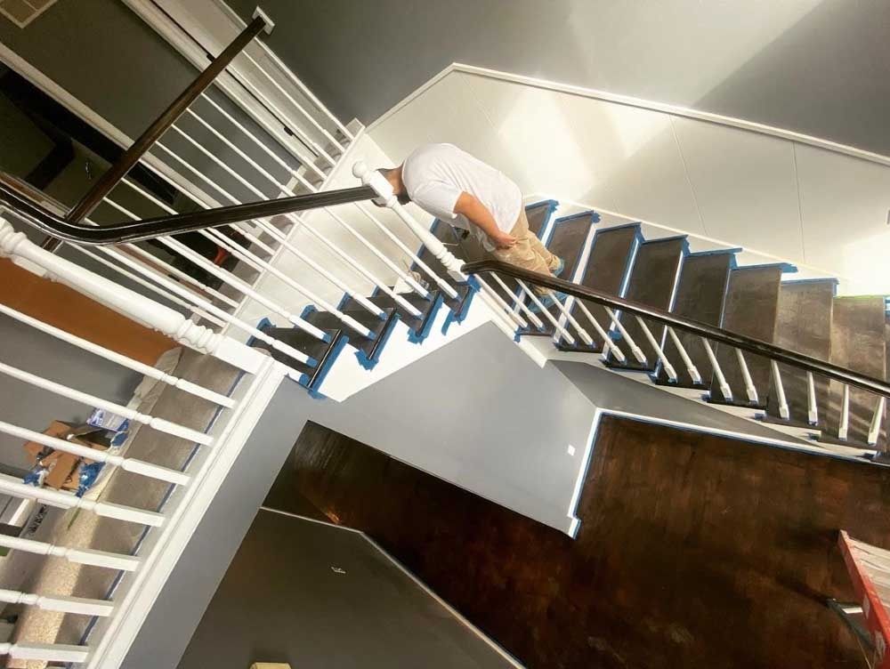 Person painting a staircase, white railings, dark wood and gray walls.