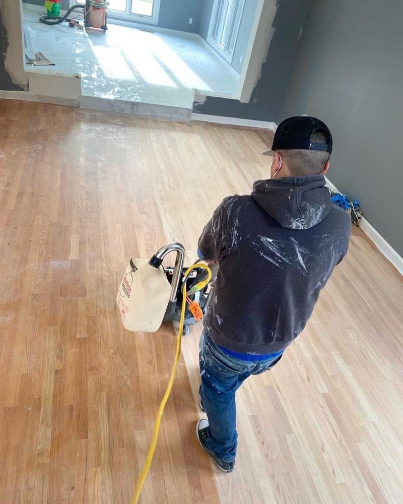 Man sanding hardwood floor, wearing a hat and jacket; interior setting with gray walls and natural light.