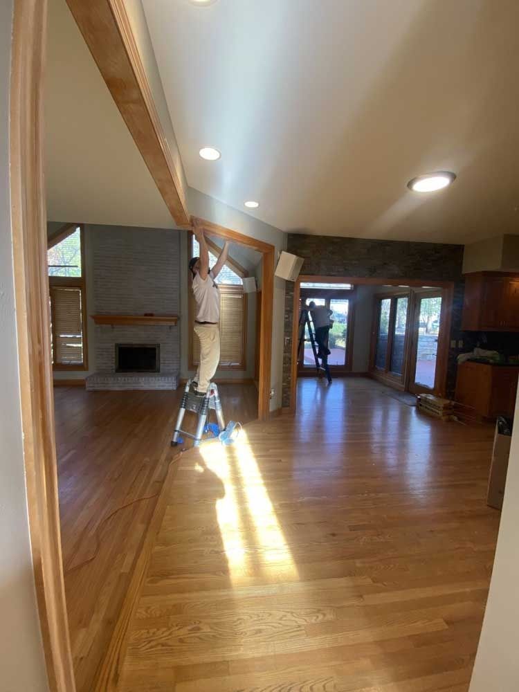 Person on ladder installing wood trim in a room with hardwood floors, a fireplace, and large windows.