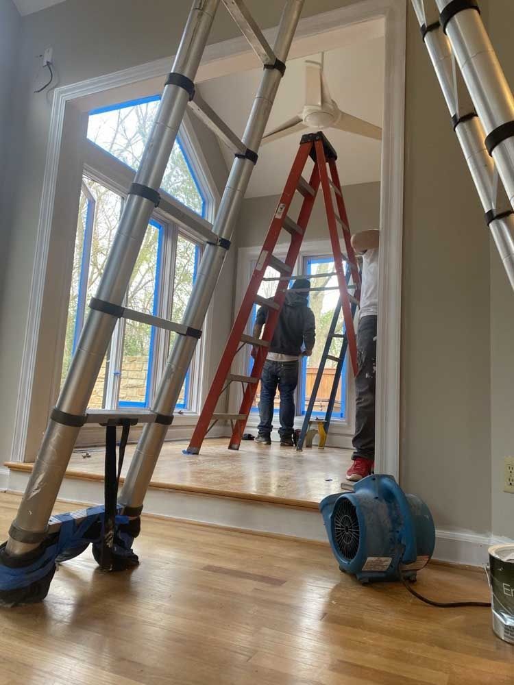 Interior view: Two people on ladders painting around a window. Blue tape covers windowpanes. A floor fan sits nearby.