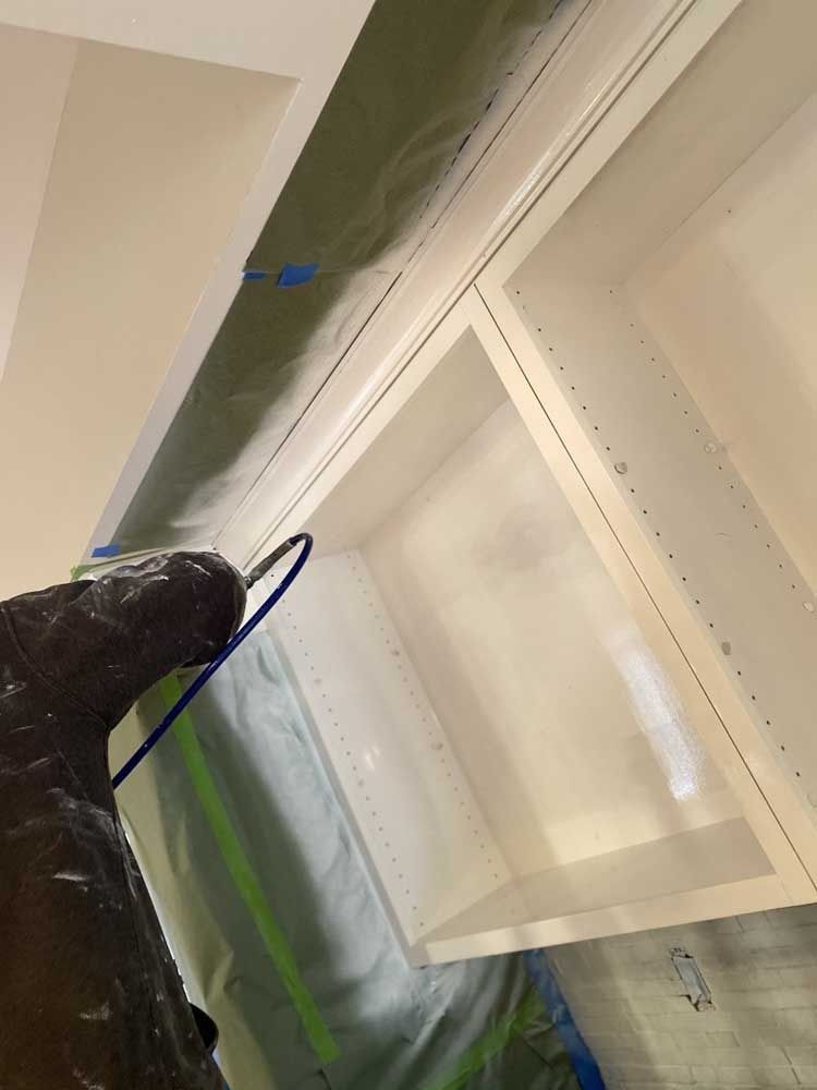 A person sprays paint onto a white cabinet in a room. Paint overspray on the walls.