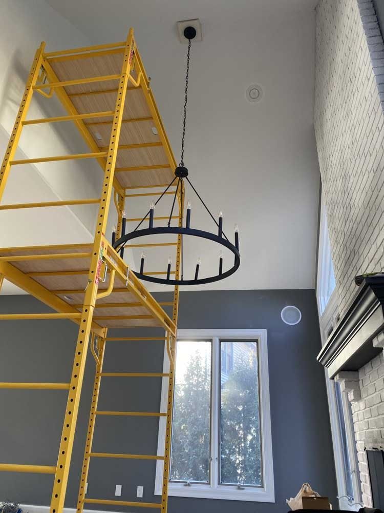 Yellow scaffolding next to a black chandelier hanging from a high white ceiling. Window and fireplace are visible.