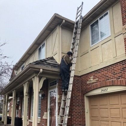 Person on a tall ladder, working on the exterior of a two-story brick and beige house. Overcast sky.