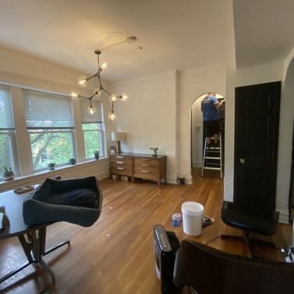 Living room with hardwood floors, a dog bed, and a worker on a ladder painting a doorway.